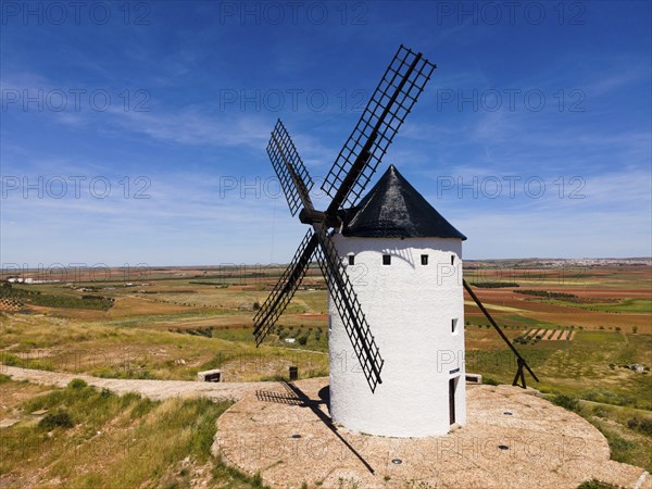 A white windmill with a black roof in front of a wide field landscape and blue sky, aerial view,
