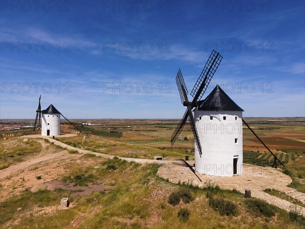 Two white windmills with black roofs in a vast landscape of fields under a sunny sky, aerial view,