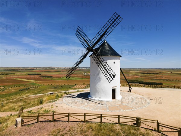 A white windmill with a black roof in the middle of fields under a clear blue sky, aerial view,