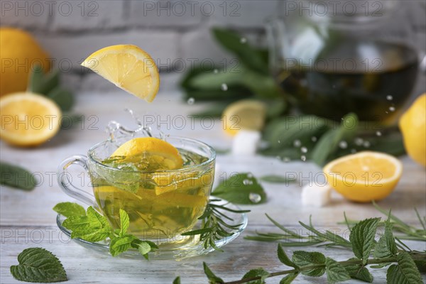 Lemon slices fall splashing into a glass of tea, surrounded by mint, sage and lemon halves