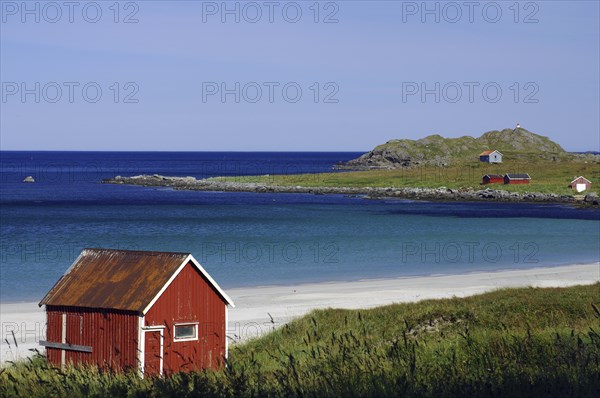 Red wooden house on a sandy beach with a view of the clear blue water and green island, Ramberg,