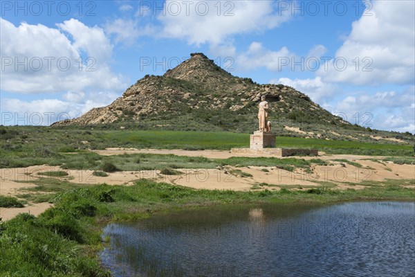 A statue stands in front of a grassy hill and a pond under a cloudy sky, Monument, Pastor