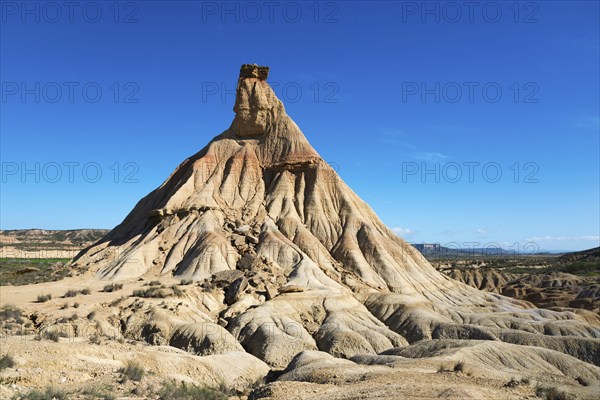 An imposing rock formation in the middle of a desert-like area under a light blue sky,