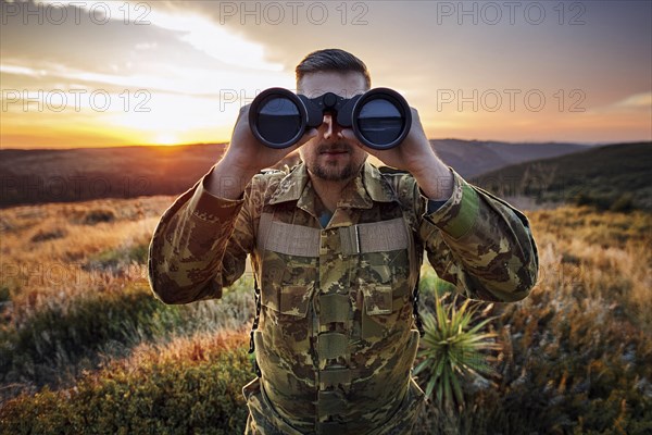Man in camouflage clothing looks with binoculars over a green landscape at sunset, AI generated, AI