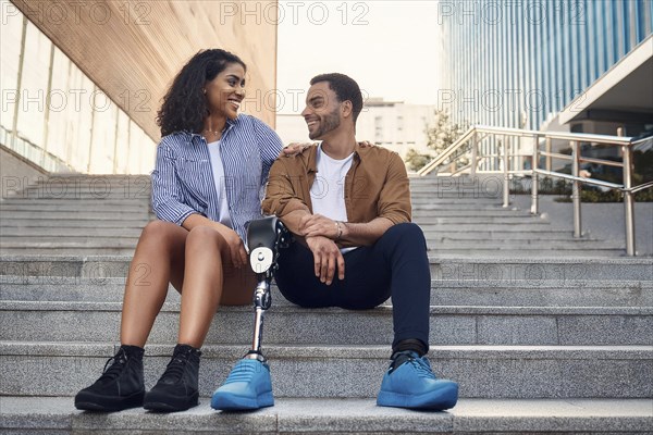 Couple, man with prosthetic leg, sitting on the steps in the city, the woman laughs at her partner