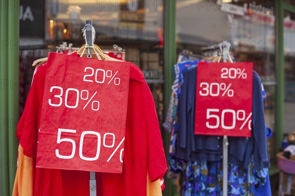 Red sign discount on a shopping bag in front of a clothing rack with t-shirts and shop window,
