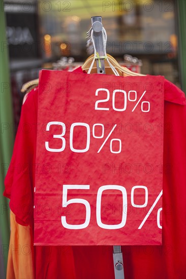 Red sign discount on a shopping bag in front of a clothing rack with t-shirts and shop window,