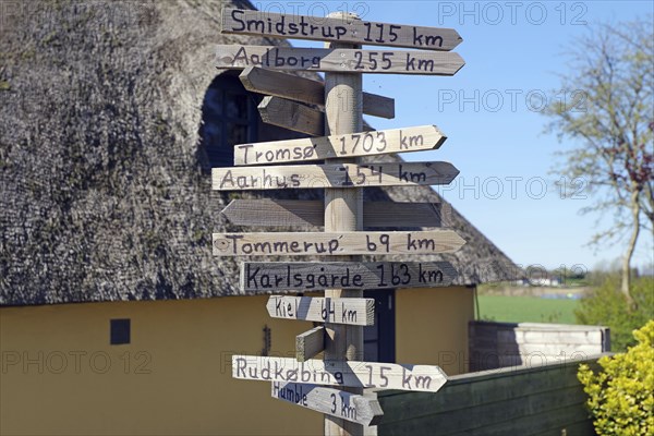 A wooden signpost indicates various distances, next to a house with a thatched roof, Langeland,