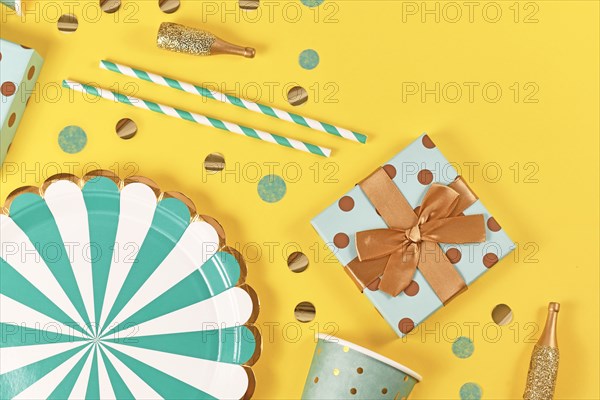 Teal blue paper plates, gift box, small golden Champagne bottles and confetti on yellow background