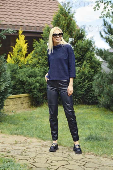 Cheerful woman in knitted blouse and leather trousers in the pathway of the garden