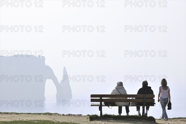 Elderly tourists sitting on bench looking at the Porte D'Aval, a natural arch in the chalk cliffs