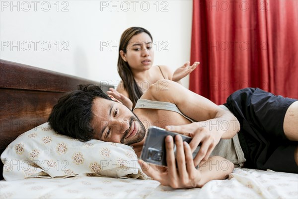 Husband with cell phone ignoring wife in bed. Annoyed woman with husband while holding cell phone