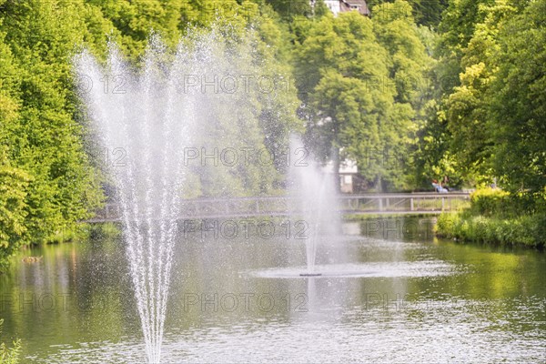 Water fountains bubble in a tranquil lake, surrounded by lush greenery and a bridge path in the