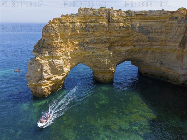 A large boat passes under natural arches made of rocks in crystal clear water on the coast, aerial