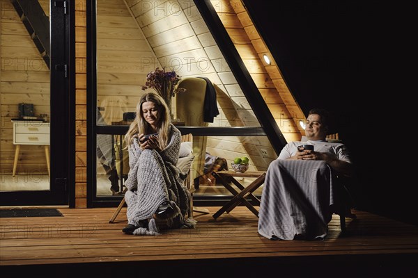 A friendly married couple is sitting on the terrace of log cabin and enjoys warm evening
