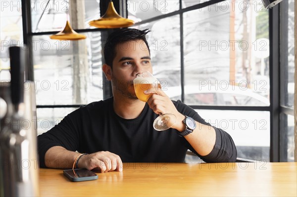 A young Latin man in a black shirt tastes a lager beer at a wooden bar table in a modern bar with