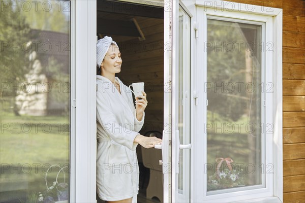 Smiling woman in bathrobe and cup of coffee in her opens the door onto terrace of wooden cabin