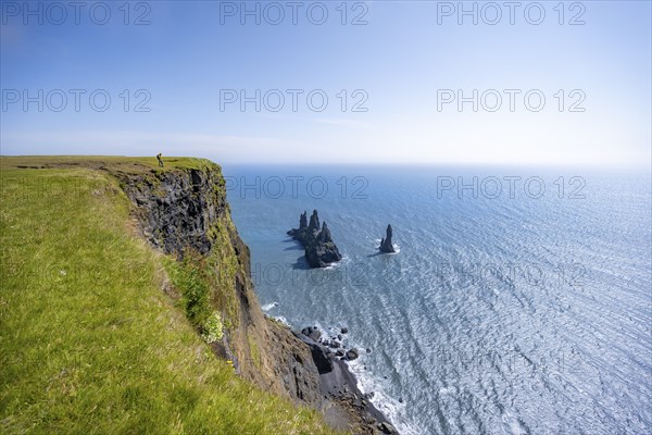 Young man standing on cliff, view from steep cliff to the rocks Reynisdrangar in the water, at