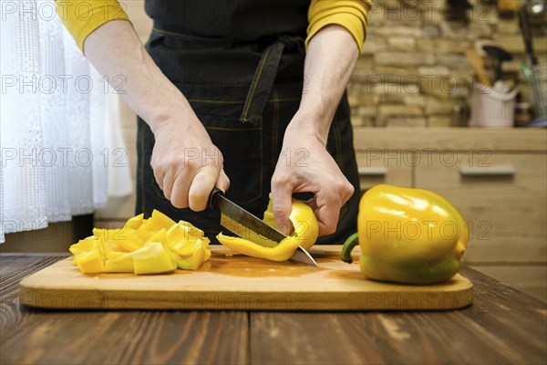 Unrecognizable man in apron slicing bell peppers on wooden cutting board in the kitchen