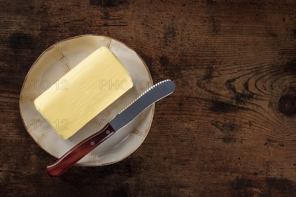 A block of butter with a knife on a plate, shot from above a dark rustic wooden background with