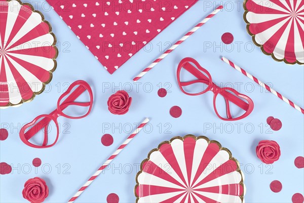 Party flat lay with paper plates, heart shaped glasses, roses, confetti and drinking straws on blue