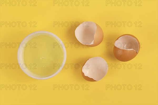 Bowl with egg white next to broken eggshells on yellow background