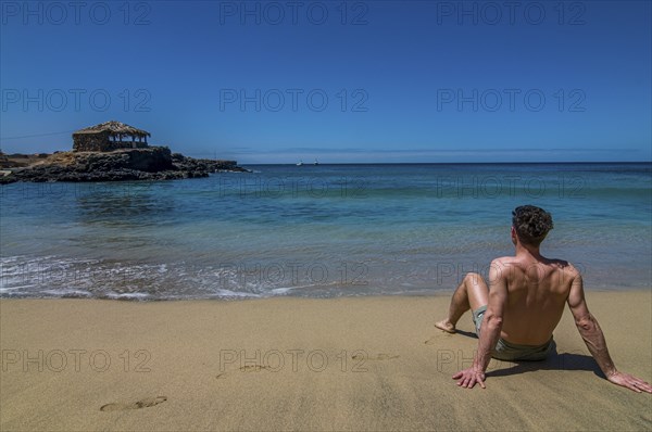 Young man with bathing-trunks, sitting on sandbeach. Tarrafal. Santiago. Cabo Verde. Africa