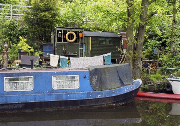 An old scruffy blue narrow boat moored next to wooden sheds with washing on the line on the