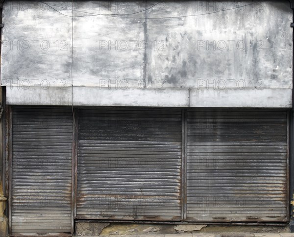 The front of an abandoned store on a street with closed dirty rusting metal shutters over the shop