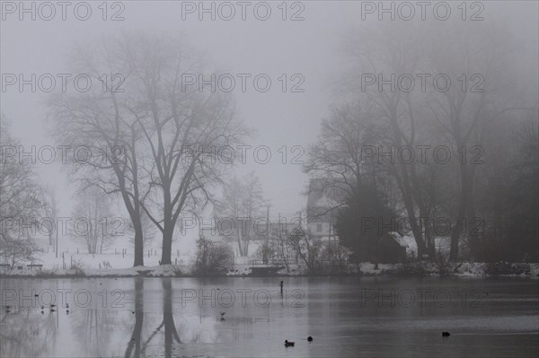 In foggy weather a lake with a house and trees in the background
