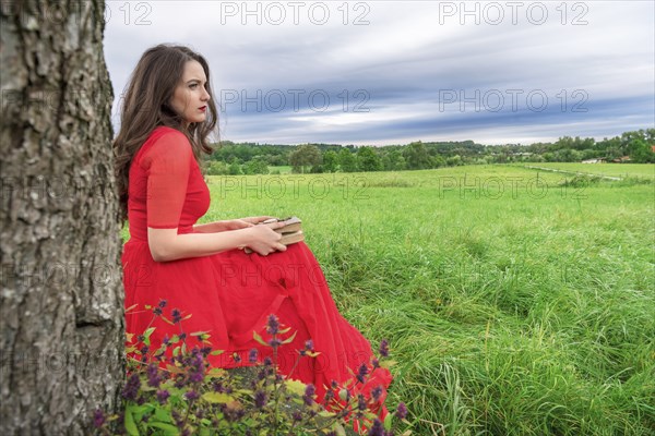 Beautiful woman, in an elegant red dress, sitting on a bench under a tree, with a book and pocket