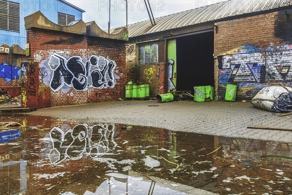 An abandoned courtyard with graffiti, puddles and green rubbish containers next to a brick wall,