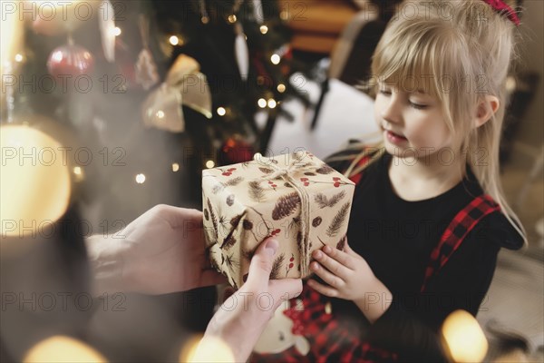 Young father in red Santa hat giving Christmas gift to his smiling daughter near decorated