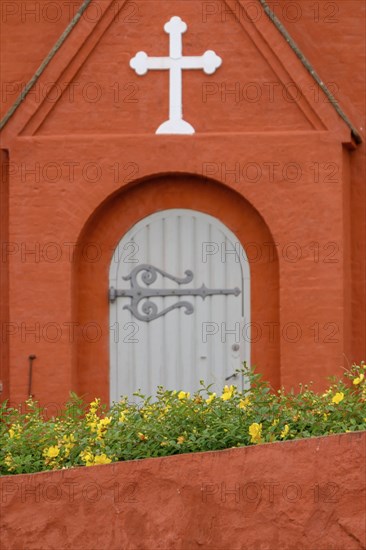 Detail of an orange-coloured church building with a white cross and Tor tor, yellow flowers in the