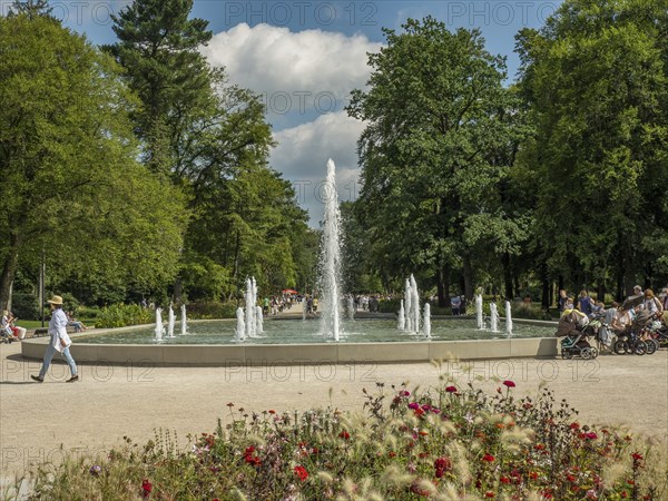 Central fountain in the park with walkers and lush greenery under a blue sky, Bad Lippspringe,