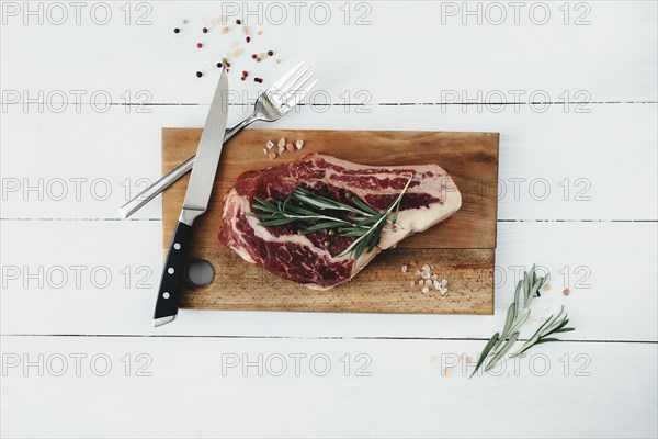 Raw ribeye steak on board, knife, fork, pepper, rosemary and pink Himalayan salt.