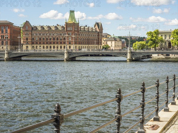 Riverside with a wrought iron railing, a bridge and historic buildings under a sunny sky,