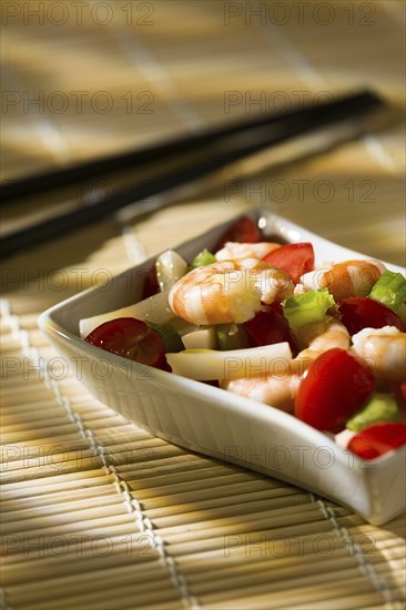 Shrimp salad with squid tomatoes celery inside a white bowl on a bamboo tablecloth and chopsticks