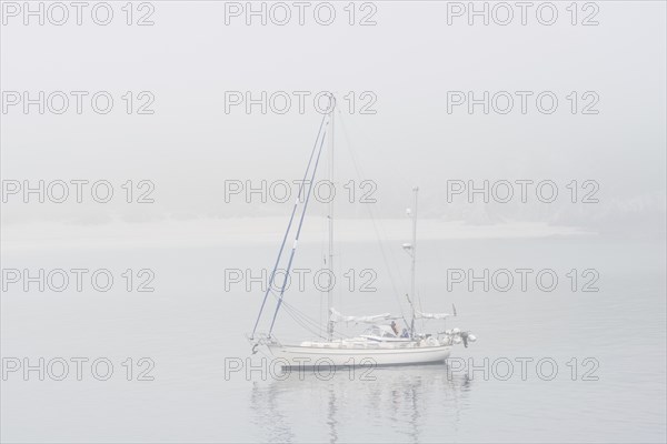 Sailboat, sailing boat, yacht with lowered sails anchored near beach during bad visibility due to