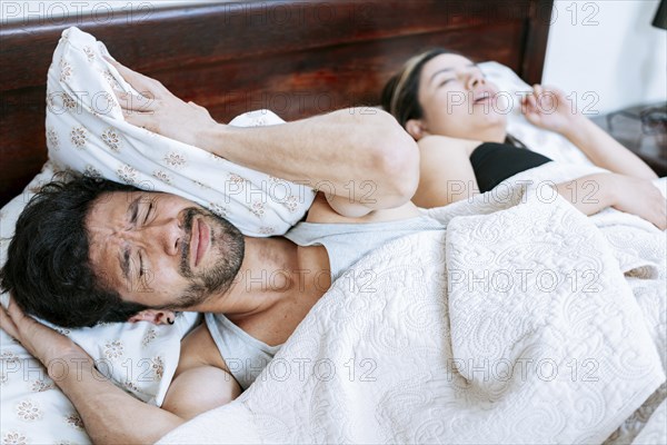Woman snoring in bedroom and husband covering his ears. Husband suffering from the snoring of his