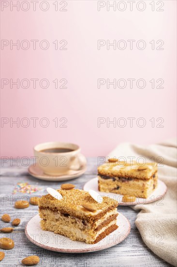 Honey cake with milk cream, caramel, almonds and a cup of coffee on a gray and pink background and