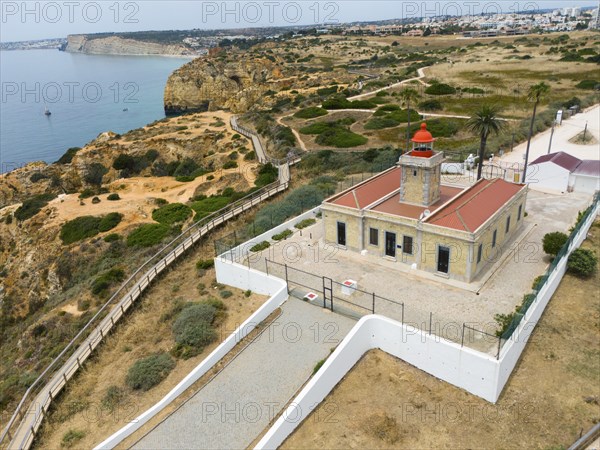 A lighthouse on the coast, surrounded by cliffs, the sea and town in the background. A path leads