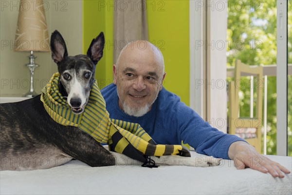 Portrait of a man posing and looking at his rescued greyhound dog with a green scarf lying on the