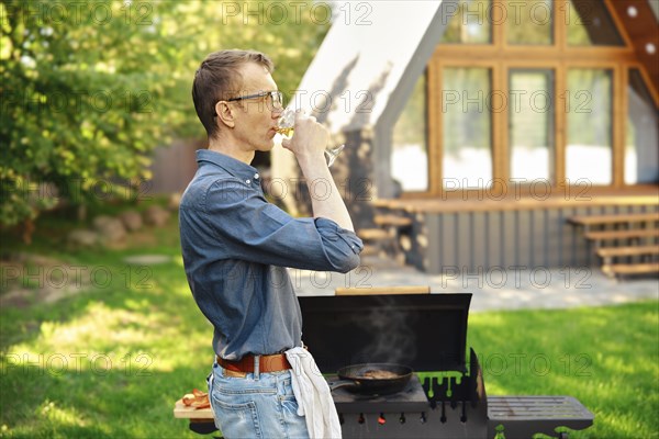 Middle-aged man drinks wine while standing in profile at the grill in the backyard of a log cabin