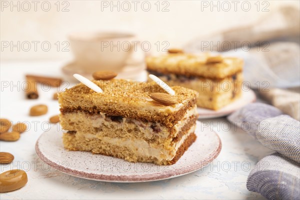 Honey cake with milk cream, caramel, almonds and a cup of coffee on a white concrete background and