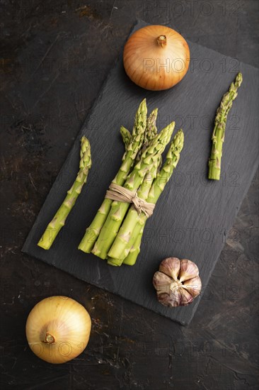 Bunch of fresh green asparagus, garlic, onion on slate board on black concrete background. Top