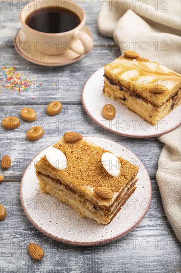 Honey cake with milk cream, caramel, almonds and a cup of coffee on a gray wooden background and