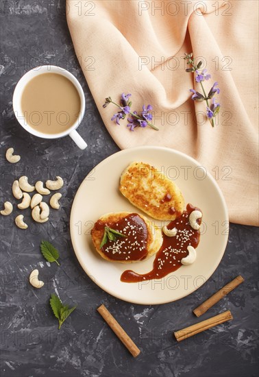 Cheese pancakes with caramel sauce on a beige ceramic plate and a cup of coffee on a black concrete