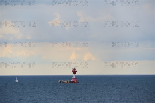 A red lighthouse stands on a calm, blue sea under a slightly cloudy sky, with a small sailing boat