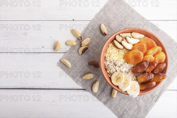 A plate with muesli, banana, dried apricots, dates, Brazil nuts on a white wooden background. top
